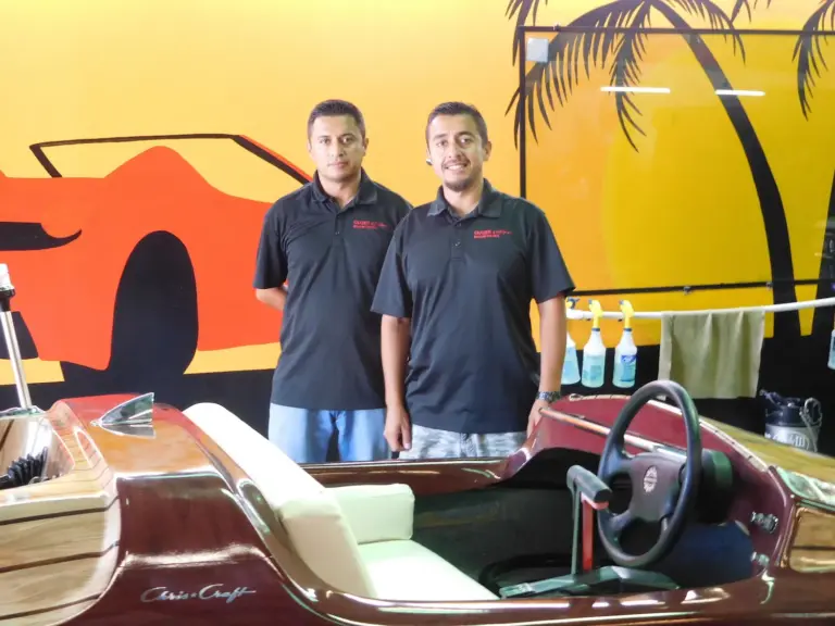 Two Shades of California team members with a classic wooden boat by the palm-tree mural